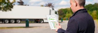 
Водитель стоит перед своим грузовым автомобилем и держит в руках планшет, на экране которого отображается форма поиска сервисных партнеров Toll Collect